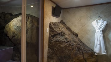 Relax in a bathroom featuring a unique rock formation, glass shower enclosure, mounted TV, and a convenient robe hanging area.