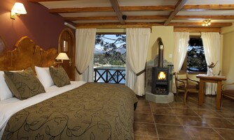 Un dormitorio con cama, cabecera de madera, mesita de noche con lámpara, chimenea de piedra, vista al exterior y una pequeña mesa con un jarrón.