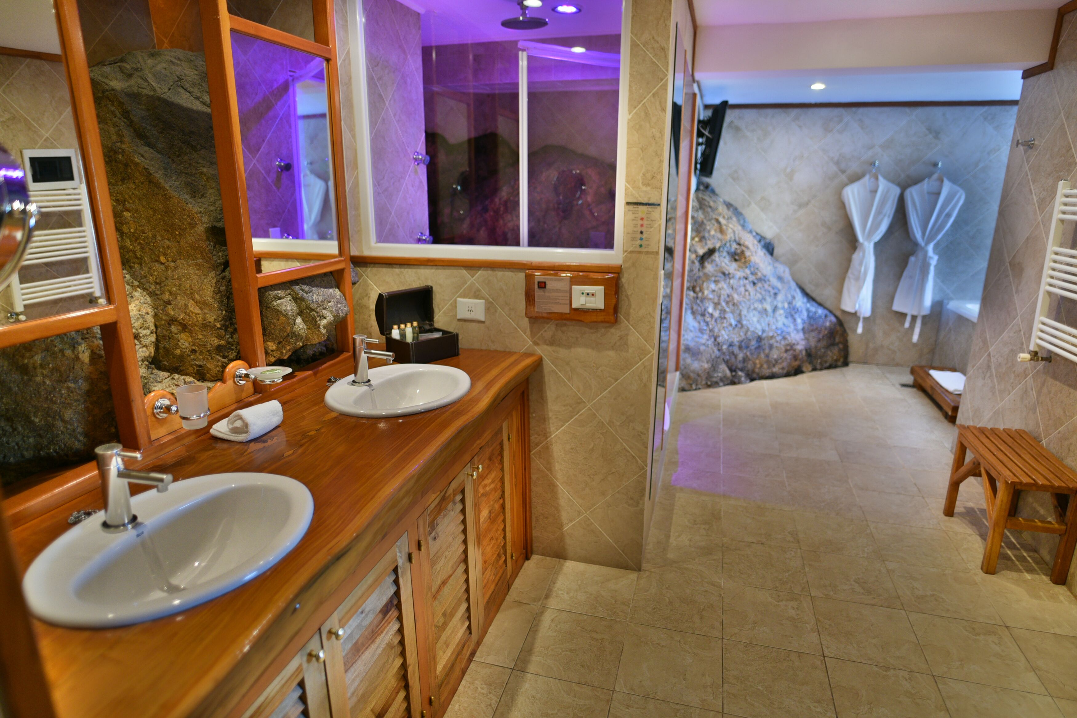 Un baño con dos lavamanos, un espejo grande, ducha con puerta de cristal y un banco de madera.