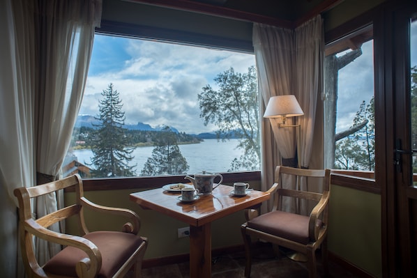 A room with two wooden chairs, a small table, a lamp, and a view of a lake and mountains through large windows. - Charming Residences & Suites with Private Spa (San Carlos de Bariloche)