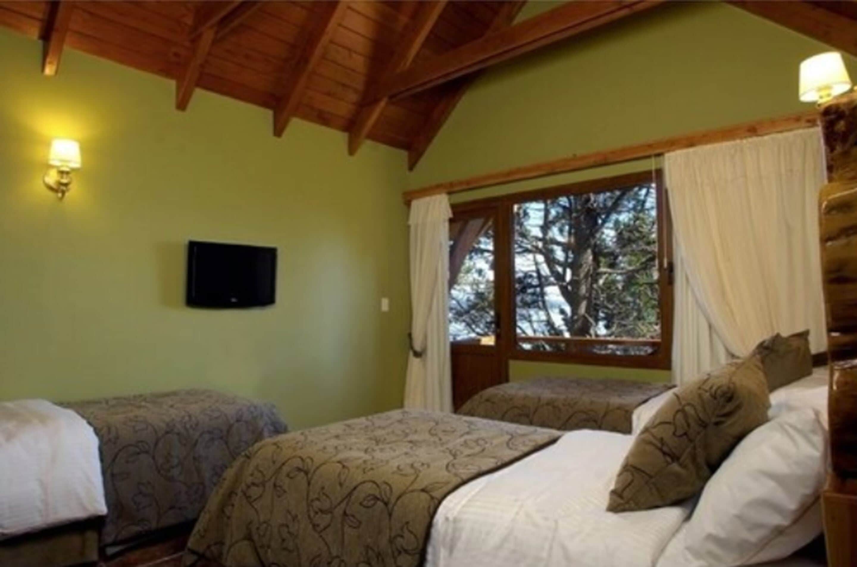 A room with two beds, a wooden ceiling, a window with curtains, and a wall-mounted TV.