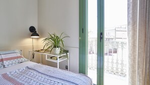 Ein Schlafzimmer mit einem Bett, einem Nachttisch mit Lampe, einer Zimmerpflanze und Blick ins Freie durch ein Fenster.