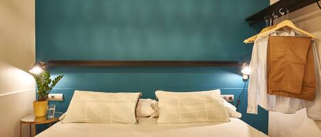 A neatly made bed with white linens, a bedside table with a plant, and a wall-mounted light.