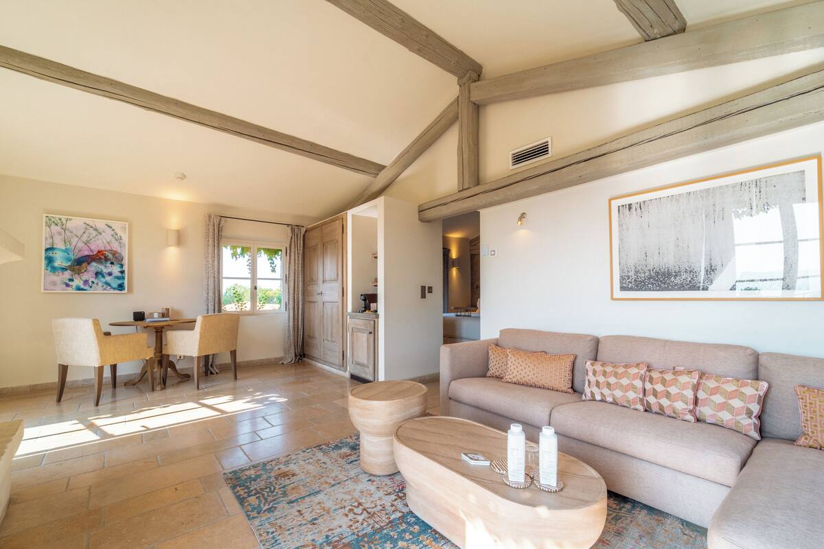 A spacious living area with a wooden ceiling, a beige sofa with patterned cushions, a round coffee table, and a dining table with chairs.
