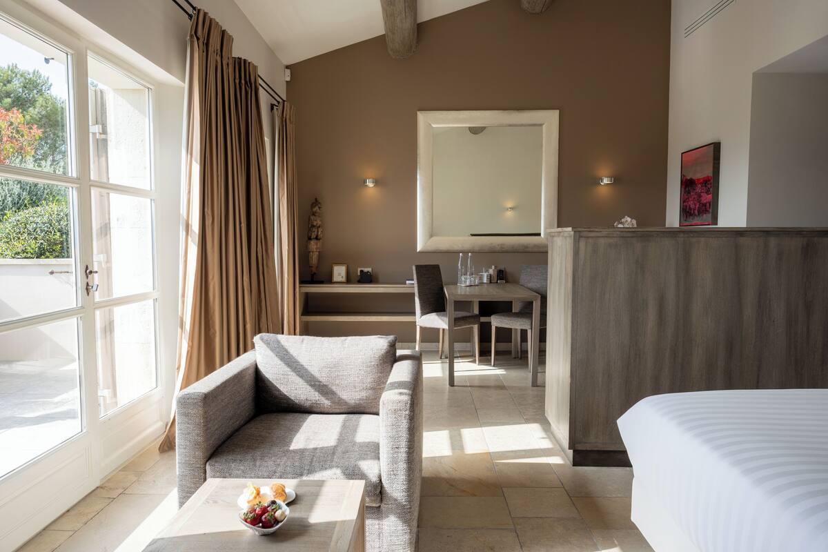 A modern hotel room with a large bed, a sofa, a dining table, and a view of the outdoors.