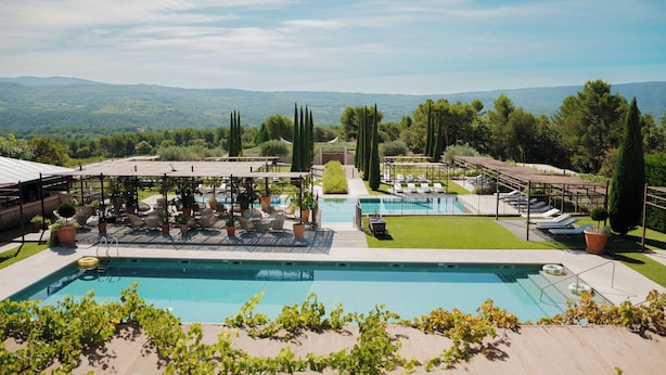 A pool area with lounge chairs and a pergola, surrounded by greenery and a view of distant hills.