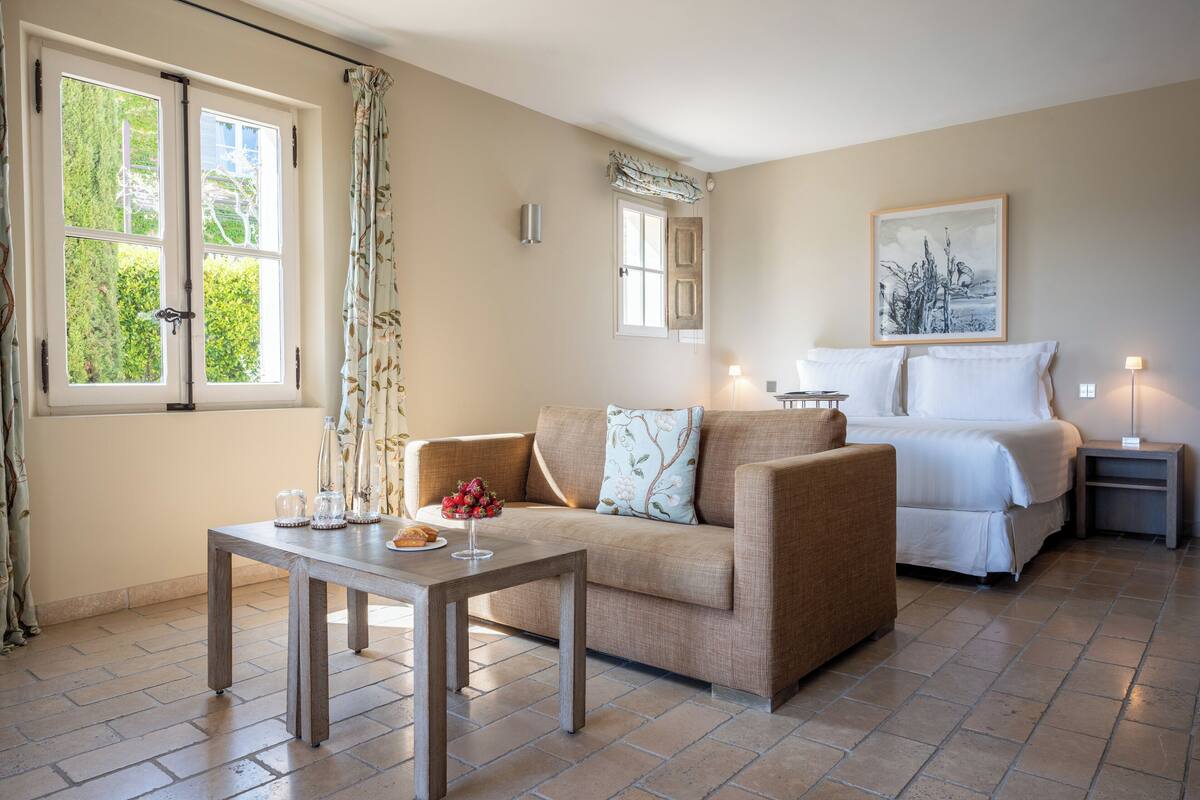 A cozy room with a sofa, a small table with a vase of flowers, a bed with white bedding, and a painting on the wall.