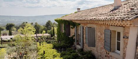 Una casa de piedra rústica con tejado de baldosas, rodeada de abundante vegetación y vistas a colinas distantes.