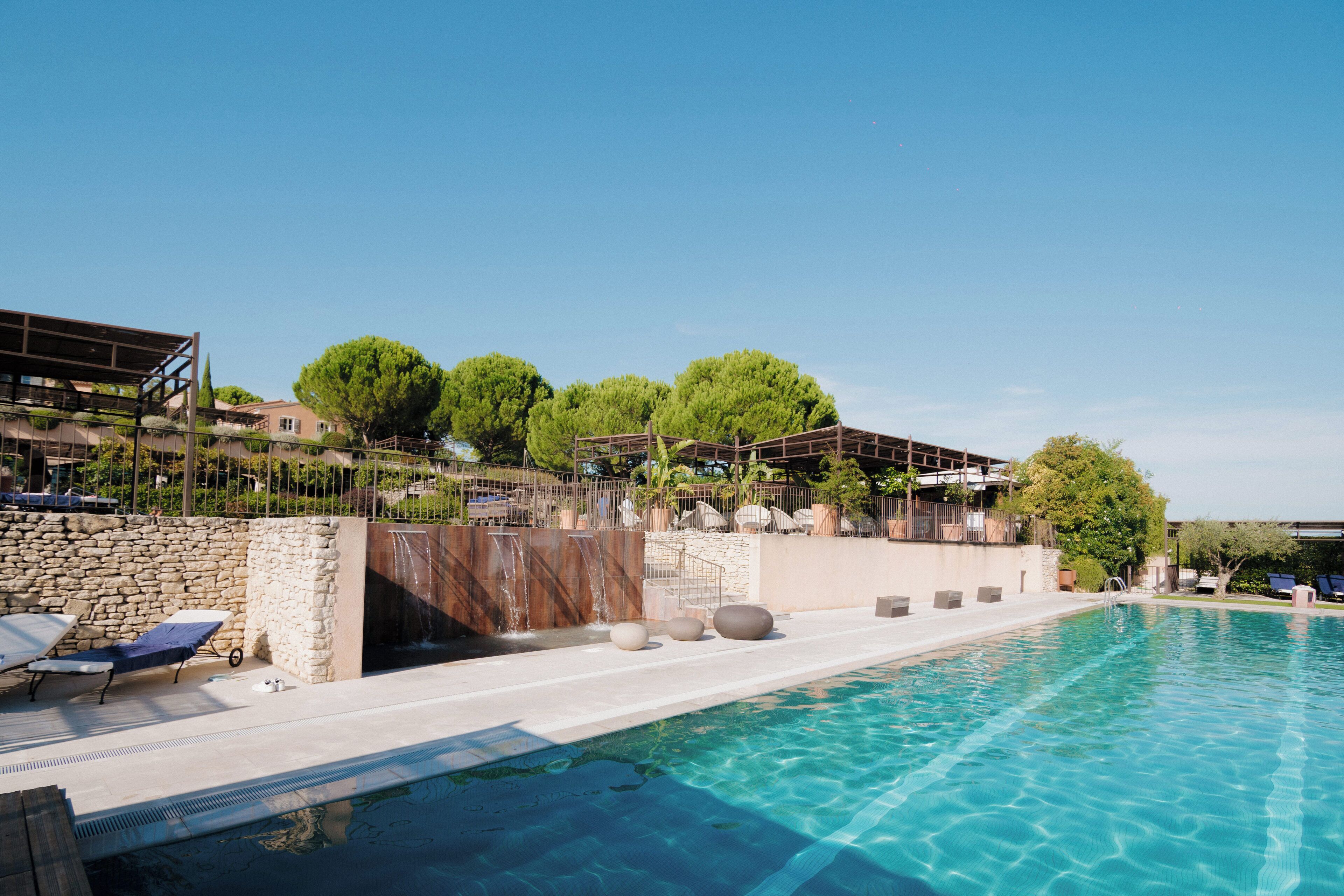 A pool area with lounge chairs, a stone wall, and a view of a building with trees.