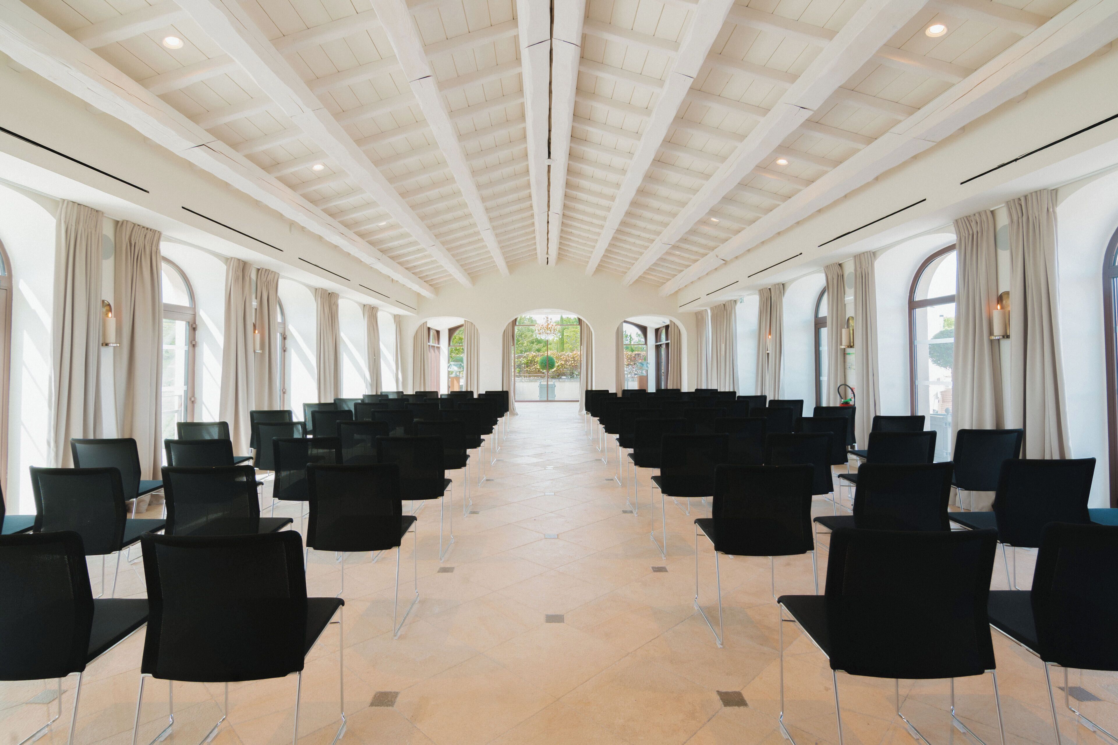 A conference room with rows of black chairs, large windows with curtains, and a high ceiling with exposed beams.