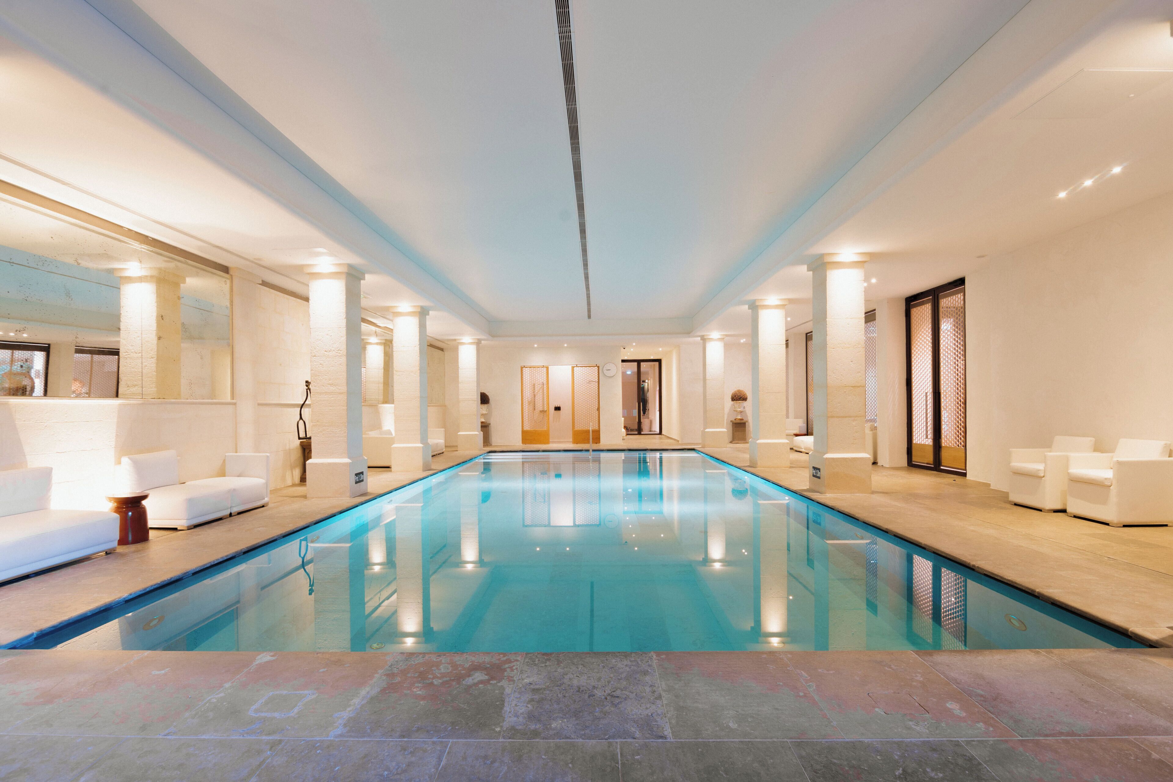 A large indoor swimming pool with a tiled floor, surrounded by white sofas and a reception area with a counter and chairs.
