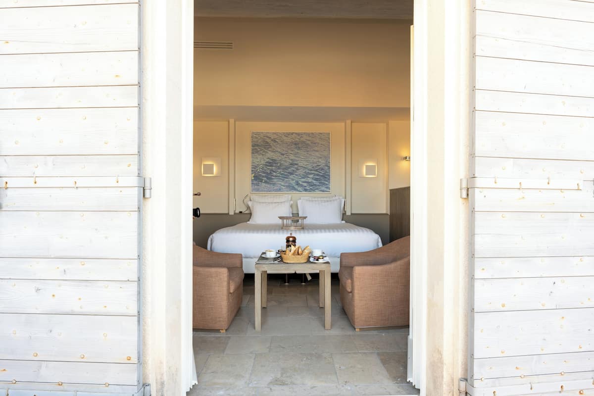 A hotel room with a bed, two armchairs, a small table with a basket of bread, and a painting on the wall.