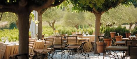 An outdoor dining area with wooden tables and chairs, surrounded by trees and potted plants.