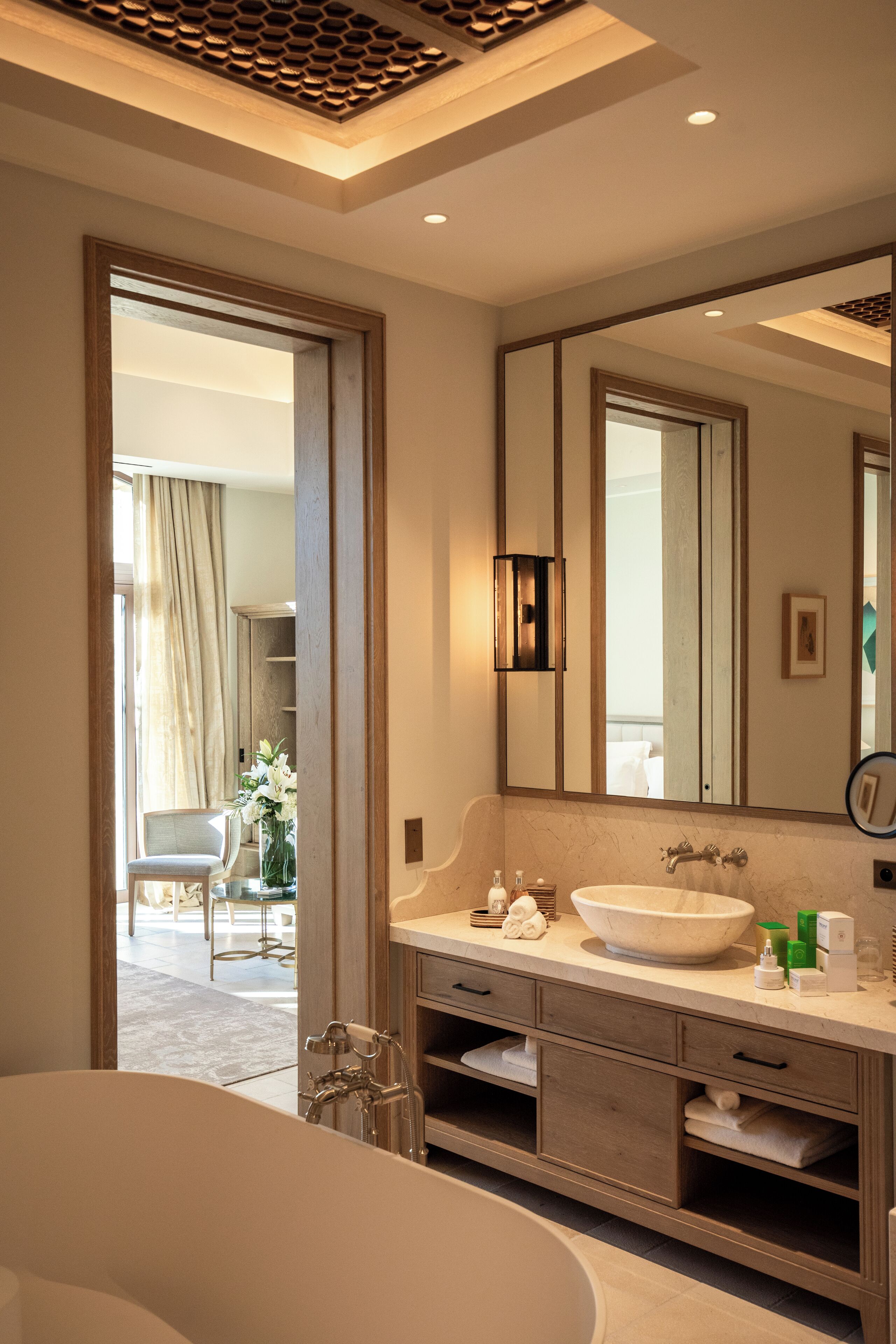 Une salle de bain moderne avec une grande baignoire, un meuble-lavabo avec un miroir et une vue sur un balcon agrémenté d’une chaise et de fleurs.