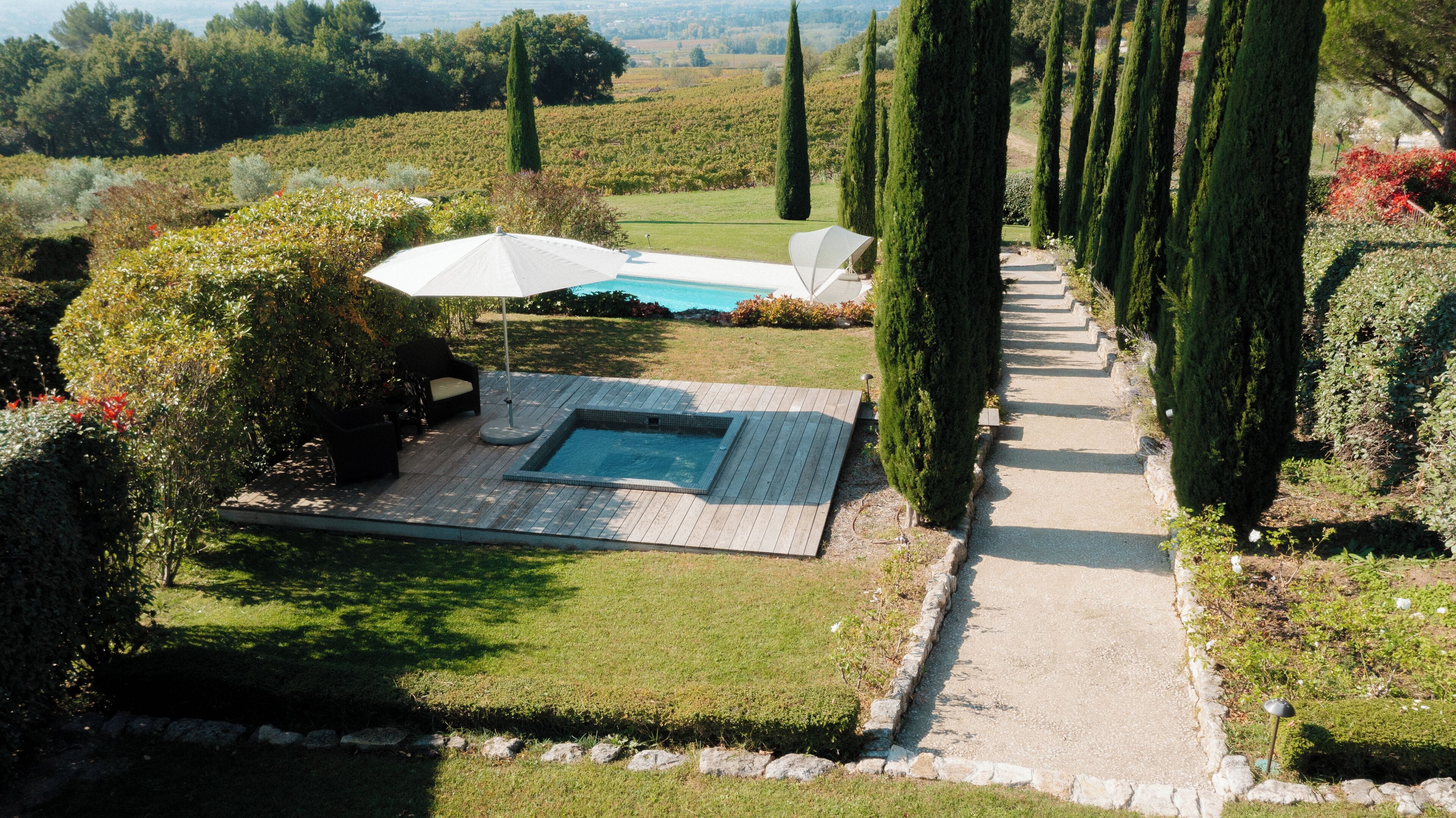 A pool area with a hot tub, surrounded by neatly trimmed hedges and tall trees.