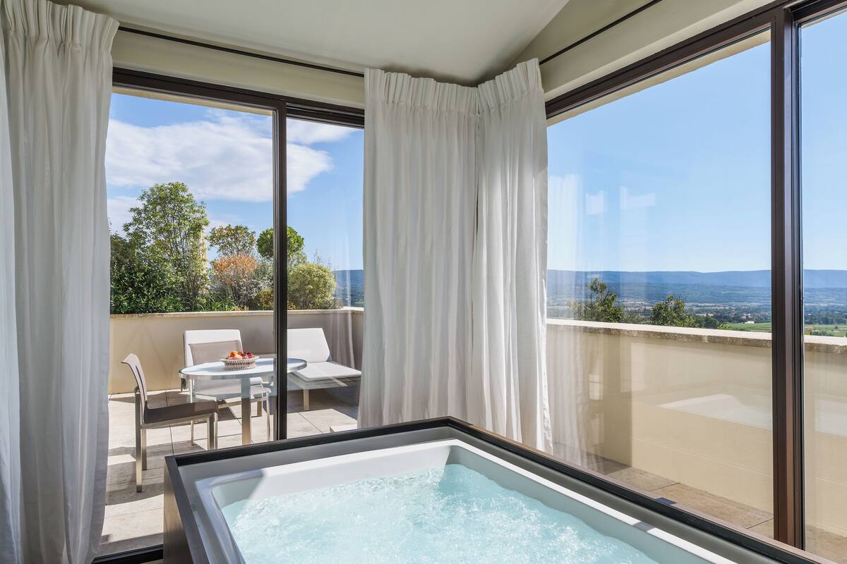 A balcony with a hot tub, white curtains, and a table with chairs.