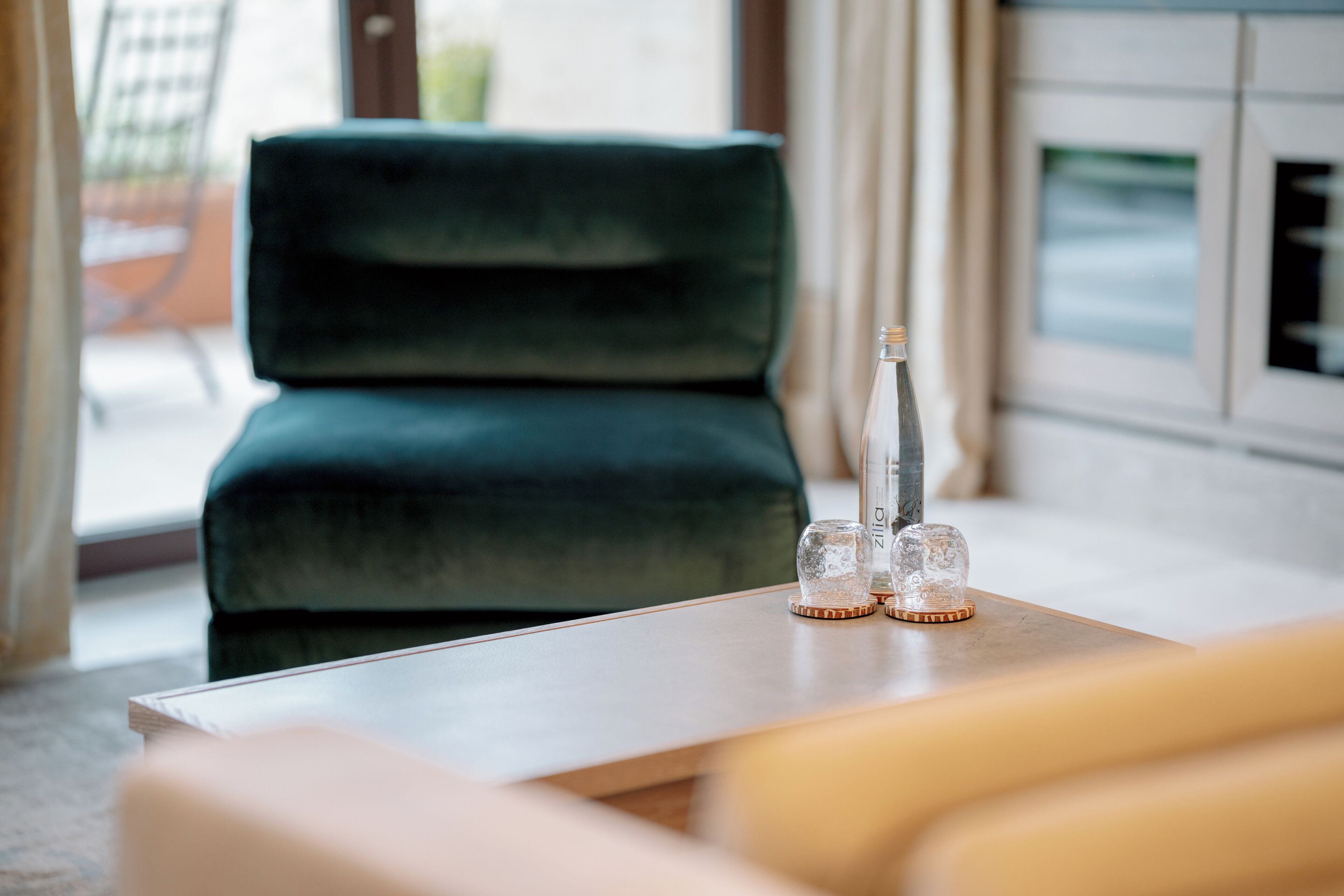 A room with a green velvet sofa, a wooden table with a bottle and glasses, and a window with curtains.