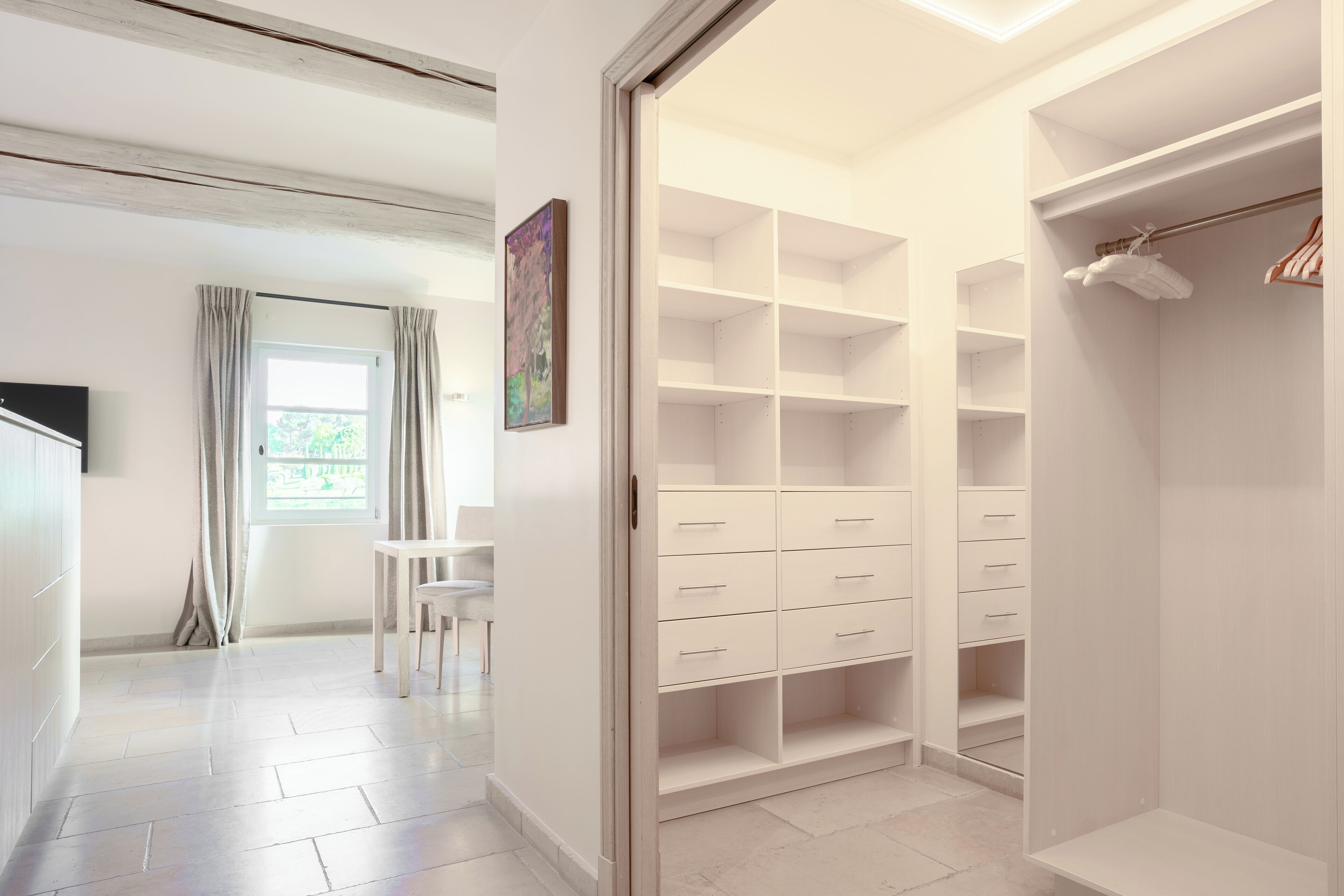 A modern, well-lit room with a large walk-in closet featuring multiple drawers and shelves, a dining area with a table and chairs, and a window with curtains.