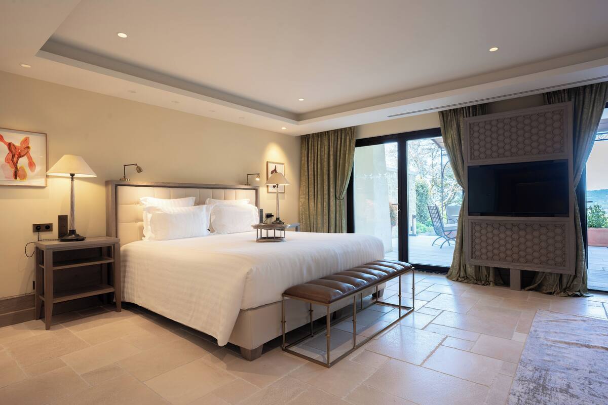 A hotel room with a large bed, a bench, a TV, and a balcony with curtains.