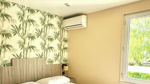 A neatly made bed with white linens and a folded towel, a wall-mounted air conditioner, and a window with a view of greenery. - Brit Hotel Essentiel Reims Taissy (Taissy)