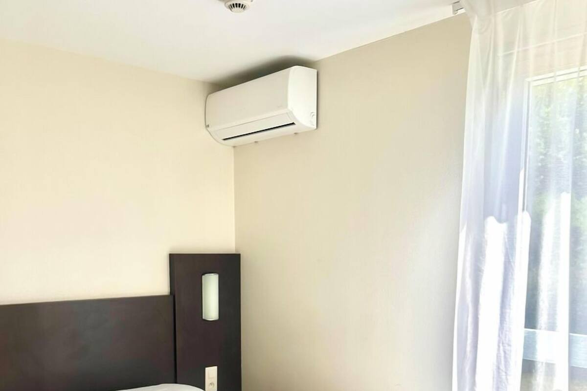 Quarto de hotel com cama, cabeceira, aparelho de ar condicionado, janela com cortinas leves e piso de madeira.