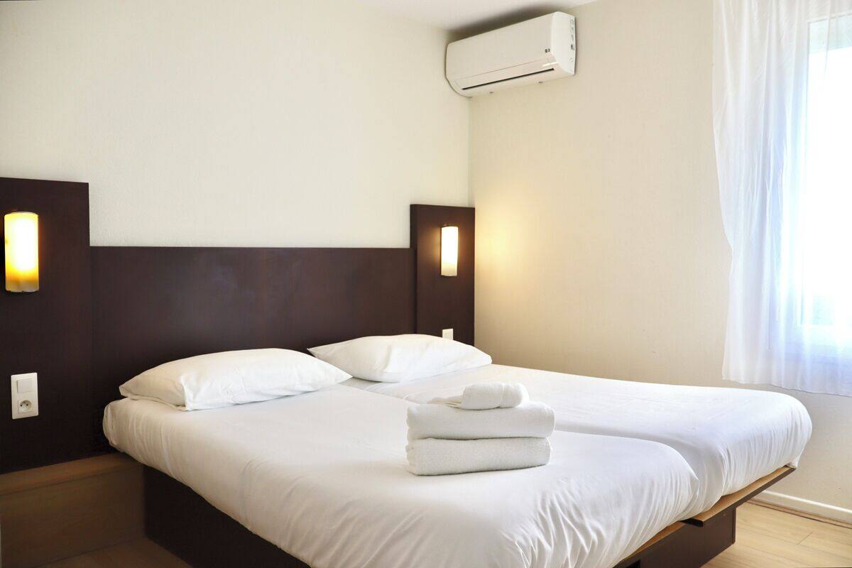 Quarto de hotel com duas camas, roupa de cama branca, cabeceira de madeira, luminárias fixadas na parede e um aparelho de ar condicionado.