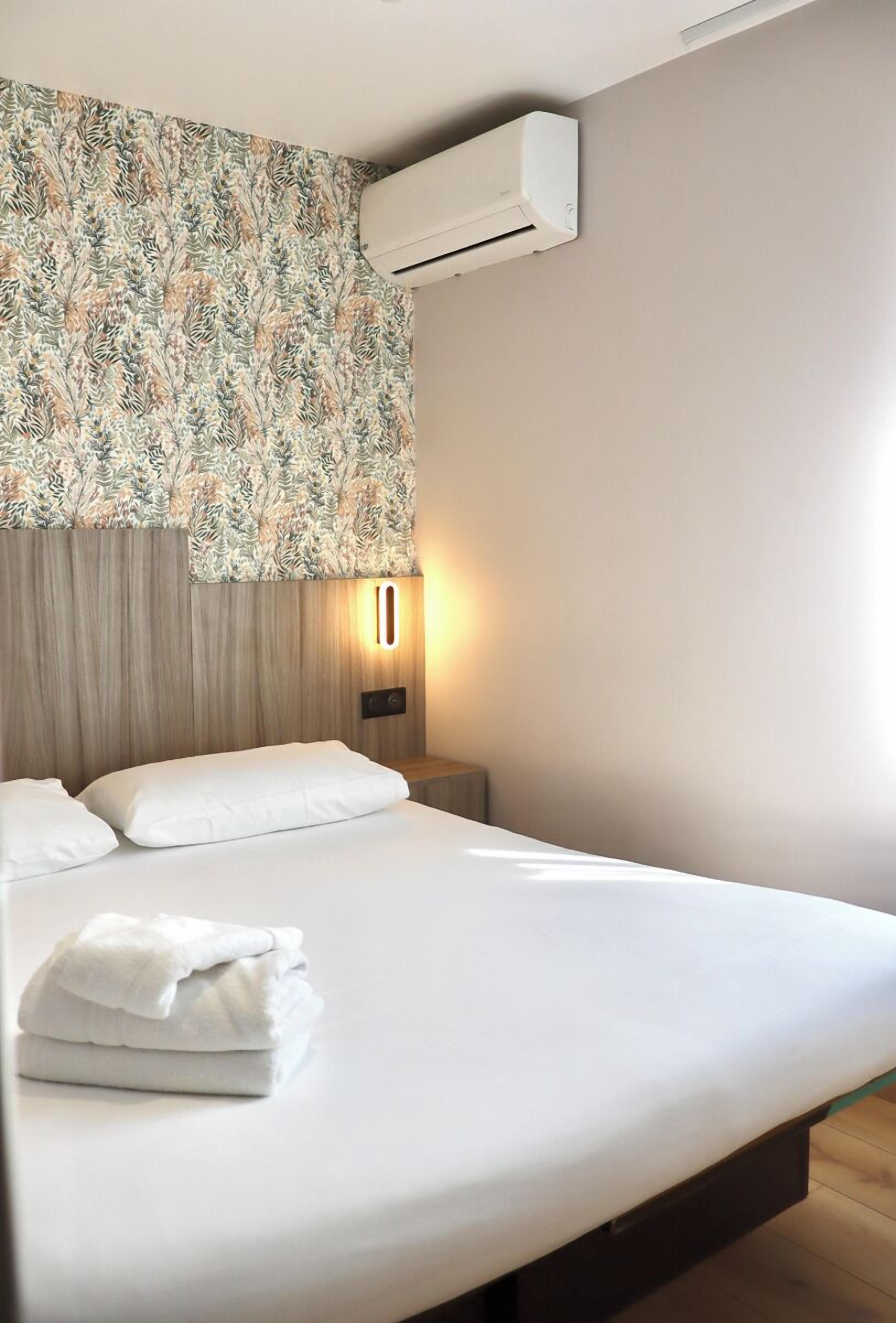 A hotel room with a bed, a wall-mounted air conditioner, a nightstand with a lamp, and a window with a floral curtain.
