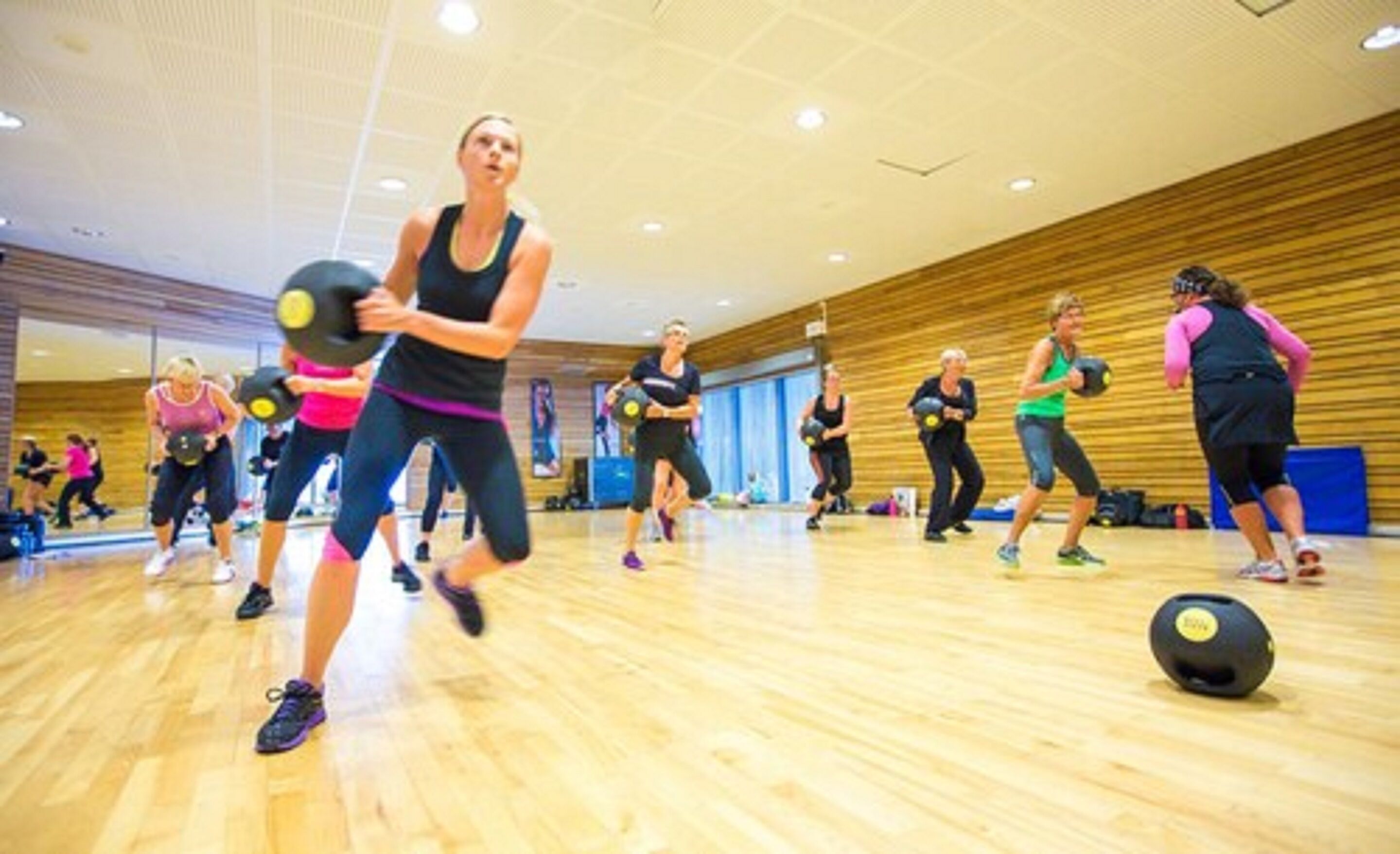 Un groupe de personnes fait de l’exercice avec des balles lestées dans une salle de sport aux murs en bois et au plafond haut.