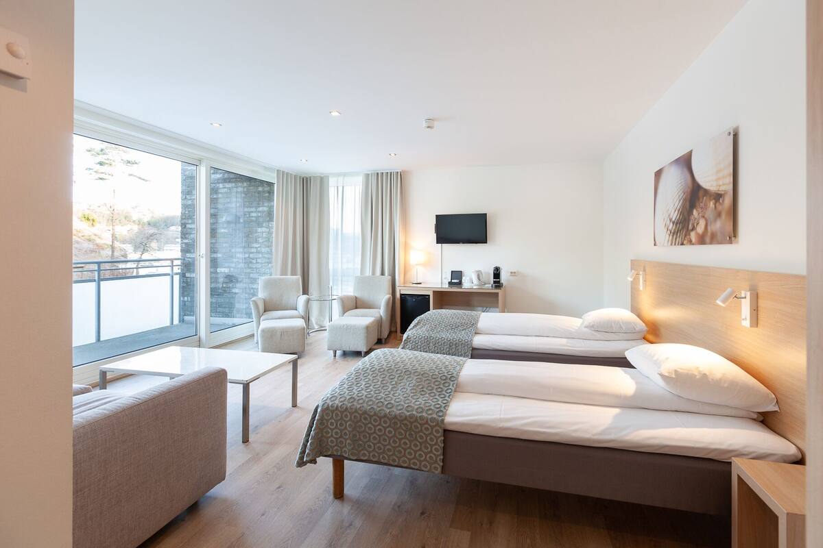 Ein modernes Hotelzimmer mit zwei Einzelbetten, einer Couch, einem Schreibtisch, einem Fernseher und Blick ins Freie.