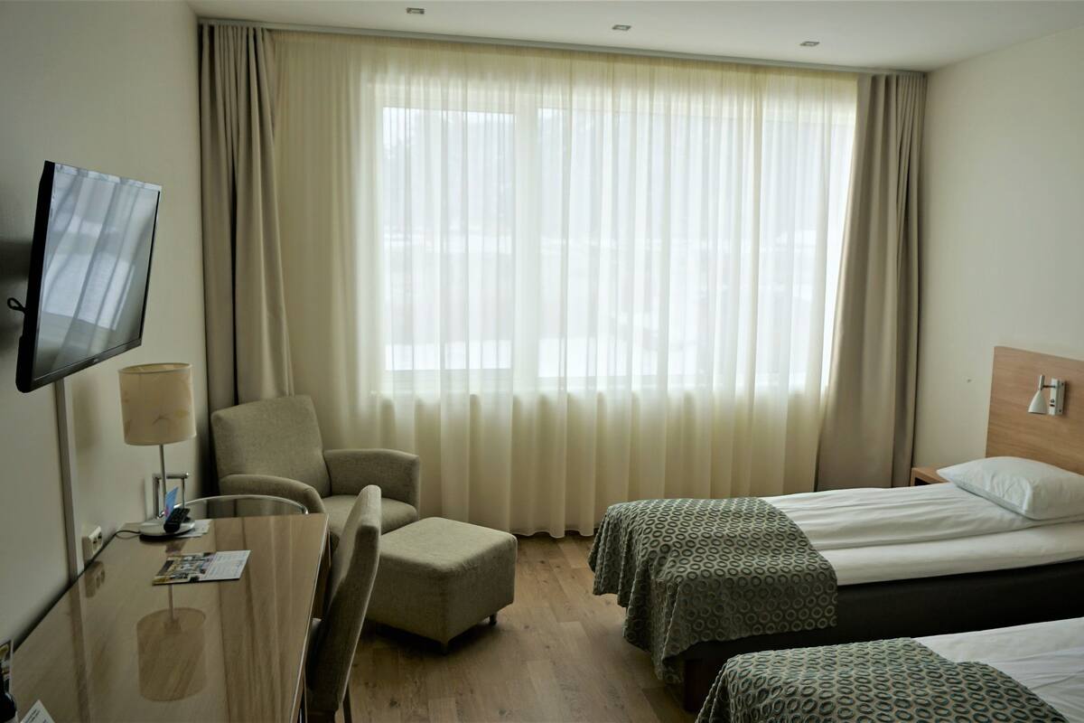 Ein Hotelzimmer mit Fernseher, Schreibtisch, zwei Betten, einem Sessel und einem großen Fenster mit Vorhängen.