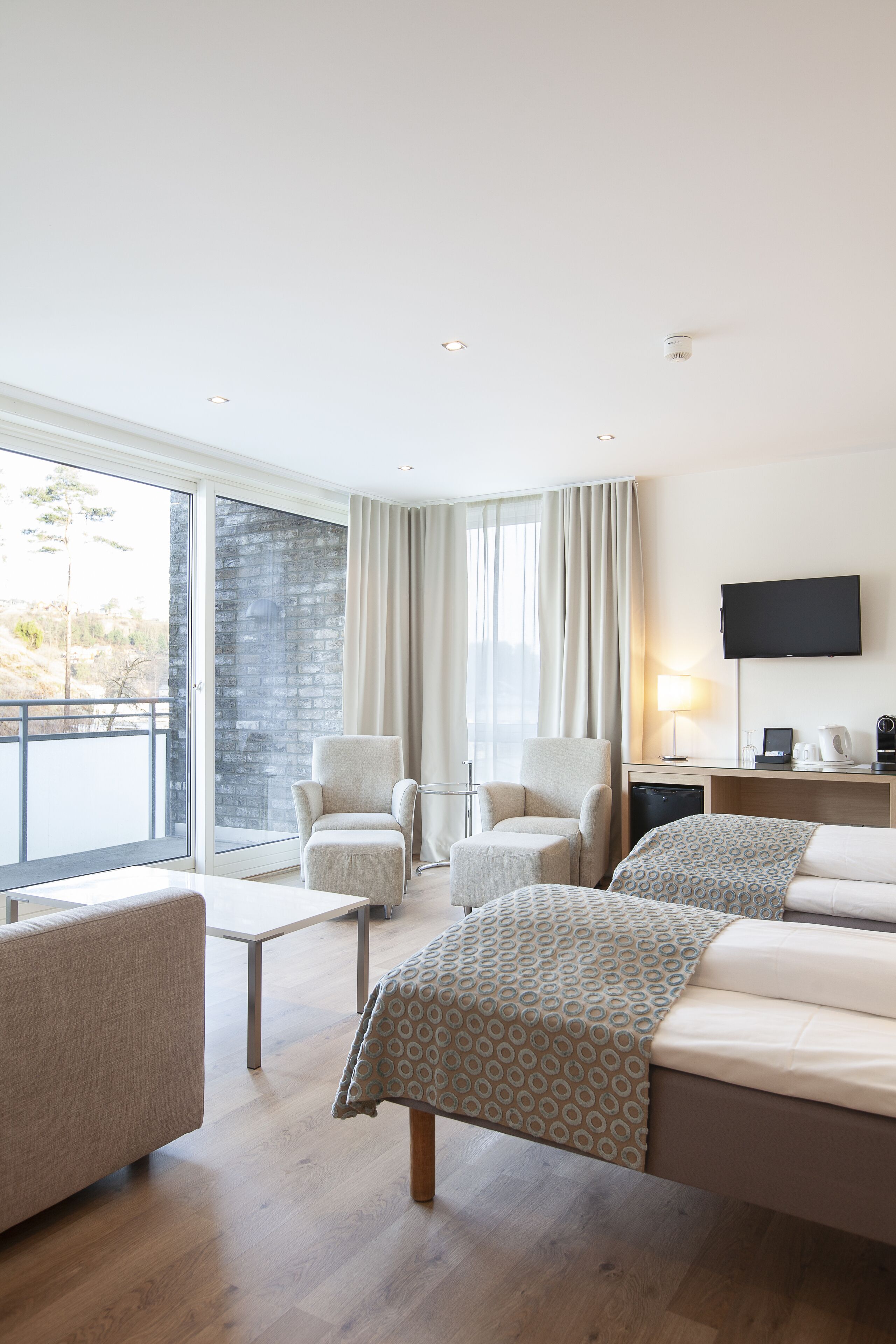 A hotel room with a bed, two armchairs, a small table, a TV, and a window with curtains.