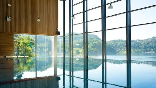 Una amplia piscina interior con paredes de vidrio transparente, que ofrece vistas a un lago y follaje circundante.