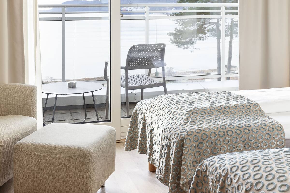 Ein Zimmer mit einer großen Schiebeglasdoor zum Balkon mit Blick auf Wasser und Bäume. Zwei Betten mit gemusterten Überwürfen, ein Sessel und ein kleiner Tisch sind vorhanden.