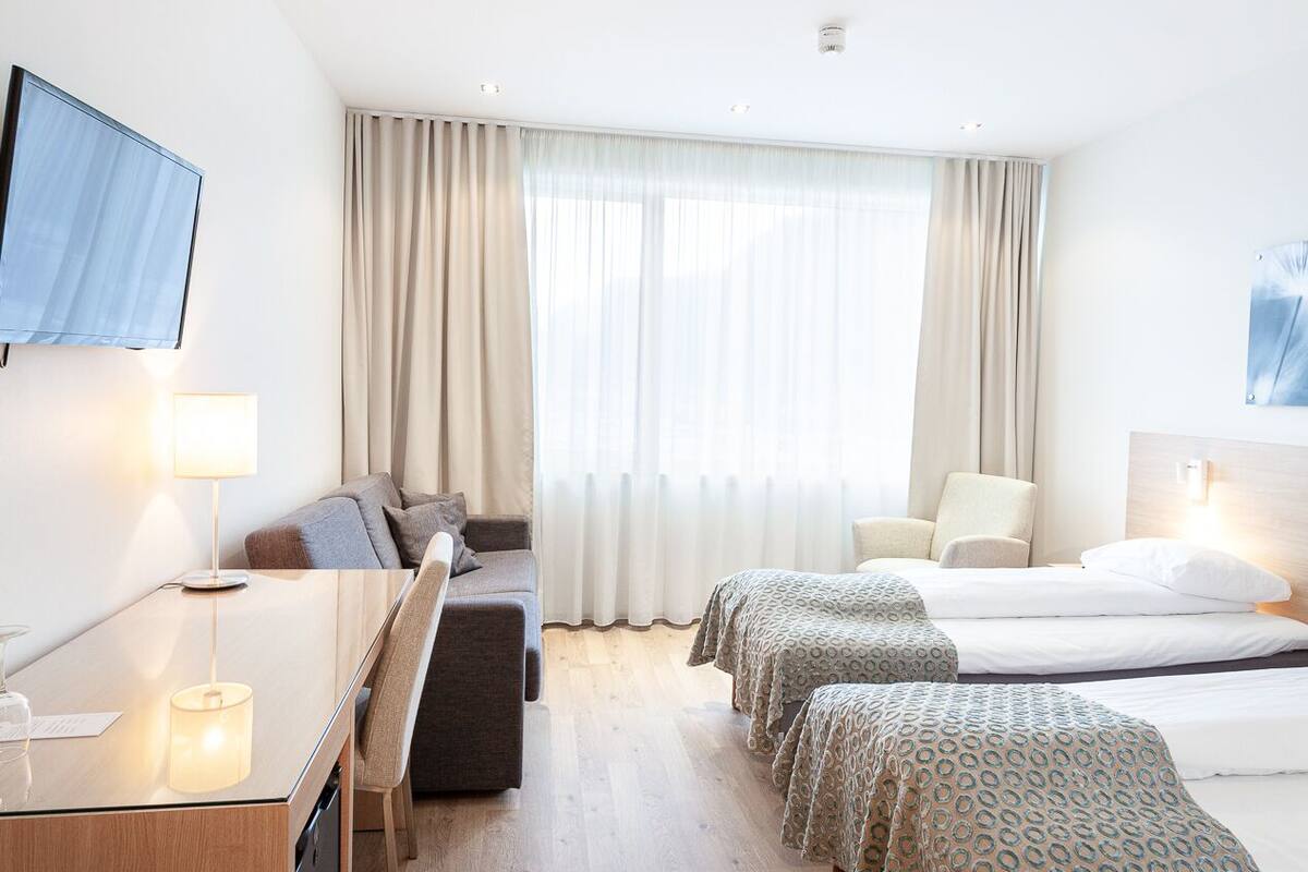 Ein Hotelzimmer mit Schreibtisch, zwei Betten, Fernseher und einem großen Fenster mit Vorhängen.