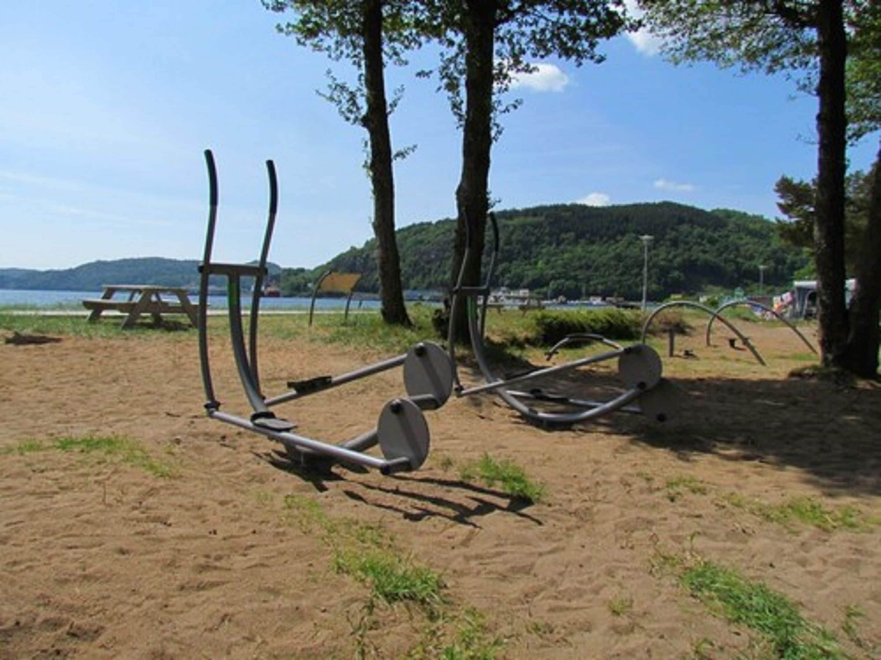 Outdoor exercise equipment on a sandy beach with trees and a picnic table in the background.