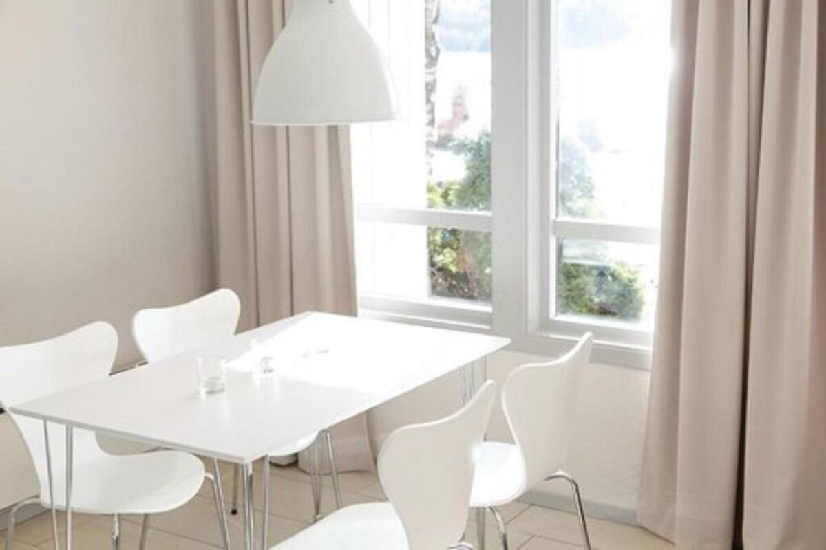 Ein moderner Essbereich mit weißem Tisch und Stühlen, großen Fenstern mit Vorhängen und einer weißen Pendelleuchte.