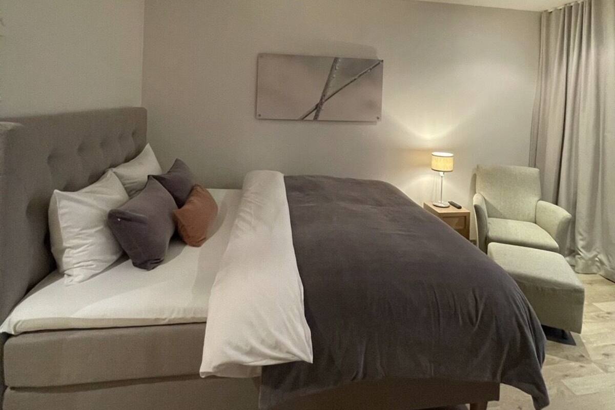 Ein modernes Hotelzimmer mit einem großen Bett, einem kleinen Sessel, einem Nachttisch mit Lampe und einem minimalistischen Wandbild.