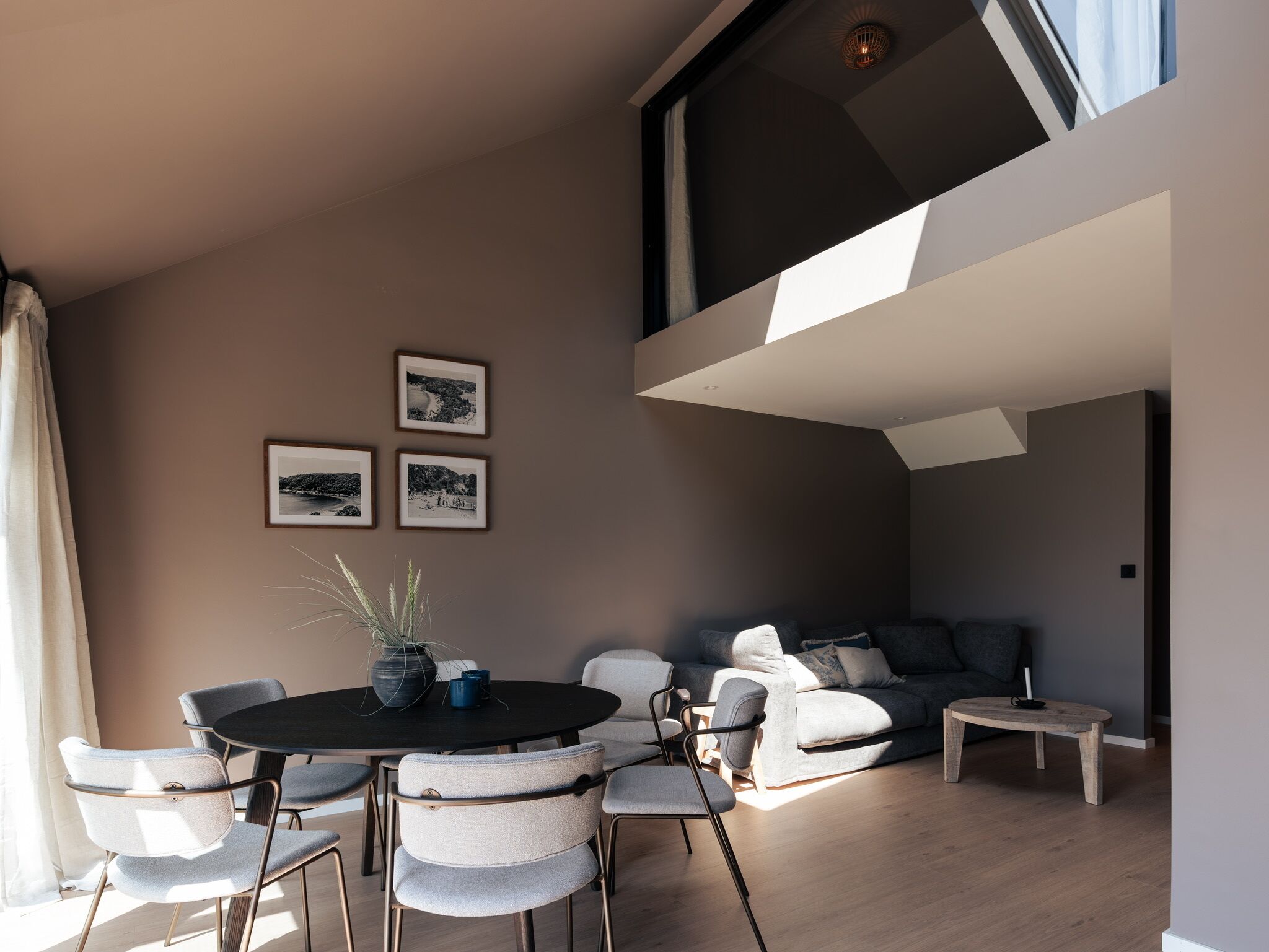 A modern living room with a round dining table, grey upholstered chairs, a sofa, and a small coffee table. There are three framed pictures on the wall.