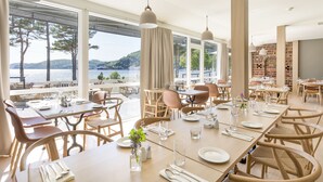 Un restaurante con mesas dispuestas para comer, ventanas amplias que ofrecen vista al agua, y una pared de ladrillo al fondo.
