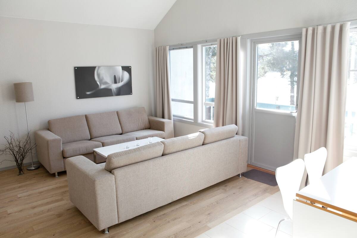 Ein modernes Wohnzimmer mit einem beigefarbenen Ecksofa, einem weißen Couchtisch und einem Essbereich mit weißen Stühlen. Eine Stehlampe und ein gerahmtes Bild sind an der Wand angebracht.