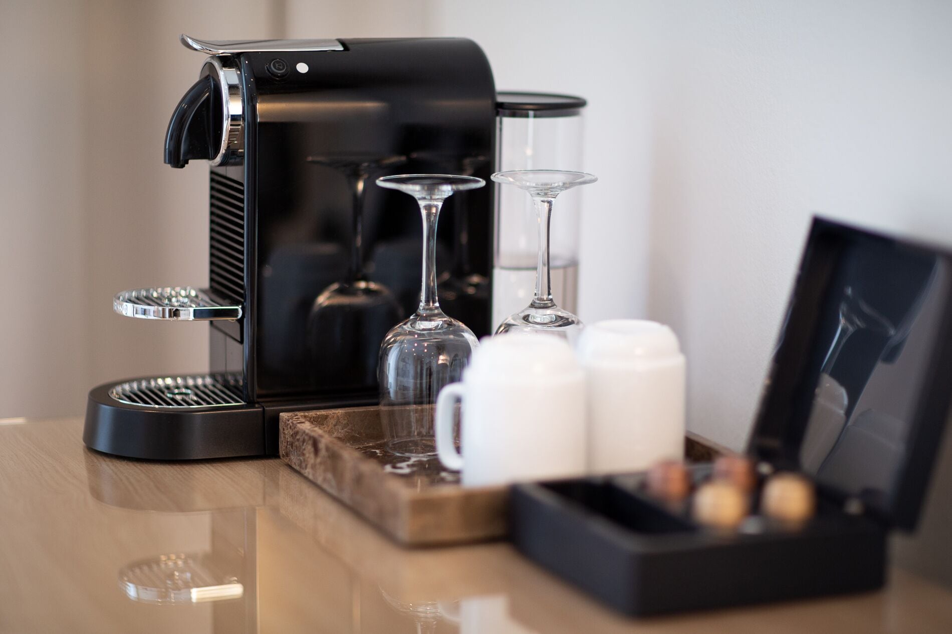 A coffee maker, two wine glasses, and a tray with cups and a box of chocolates on a wooden table.