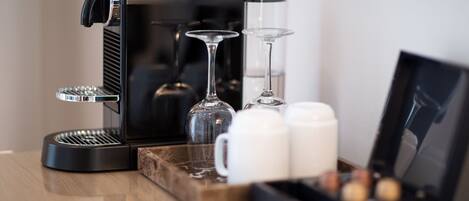 A coffee maker, two wine glasses, and a tray with cups and a box of chocolates on a wooden table.