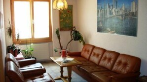 Ein Wohnzimmer mit braunem Ledersofa, einem Holztisch und einem Stadtbild. Zwei Fenster mit Vorhängen, eine Topfpflanze und eine Lampe auf dem Tisch sind zu sehen.