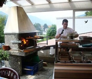 Un cocinero preparando comida al aire libre, con un horno de piedra y diversos utensilios e ingredientes sobre la mesa.