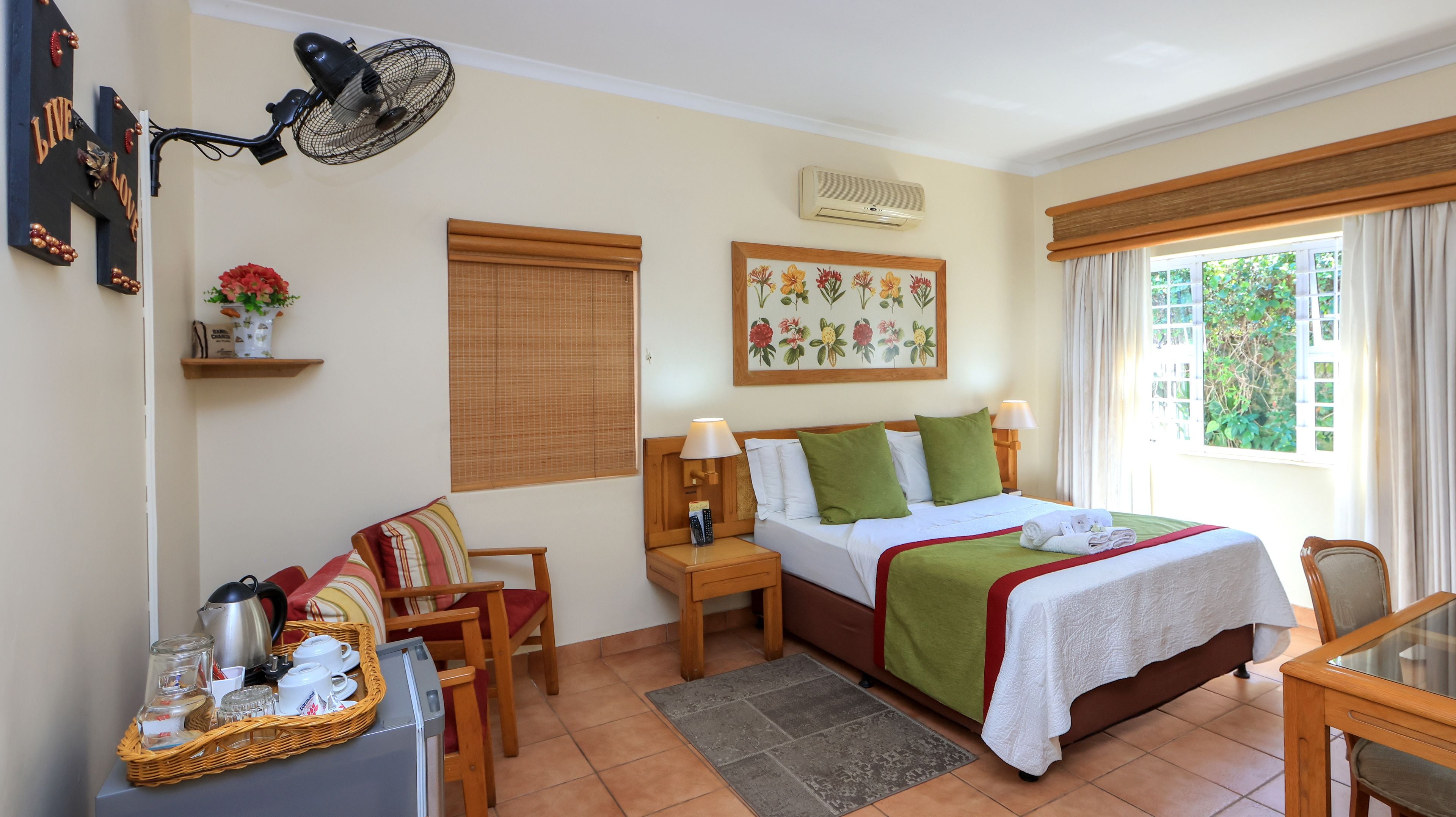 A hotel room with a bed, bedside tables, a desk with a chair, a fan, a window with curtains, and a wall-mounted air conditioner.
