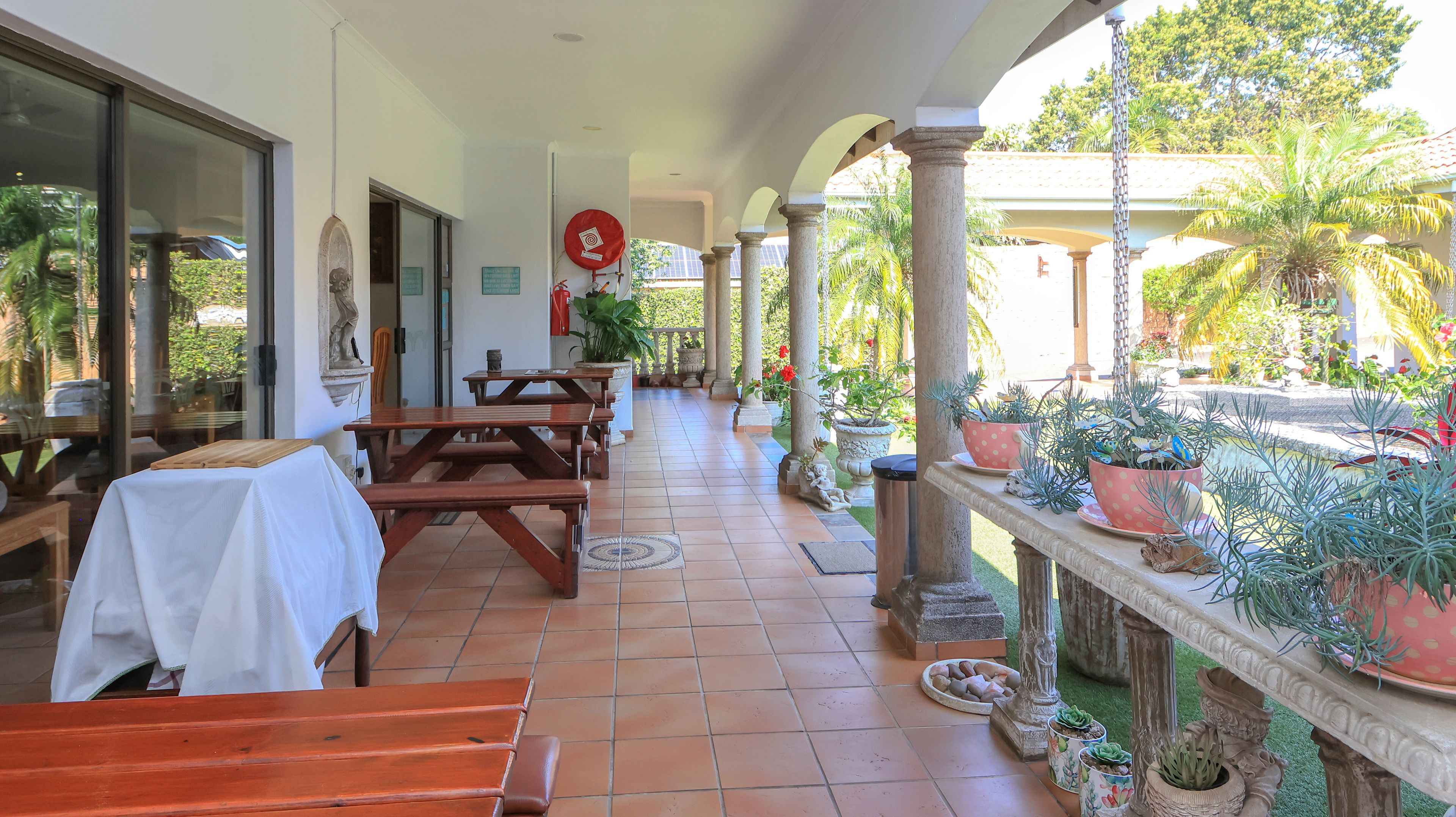 A covered outdoor seating area with wooden tables and benches, potted plants, and a view of a garden.