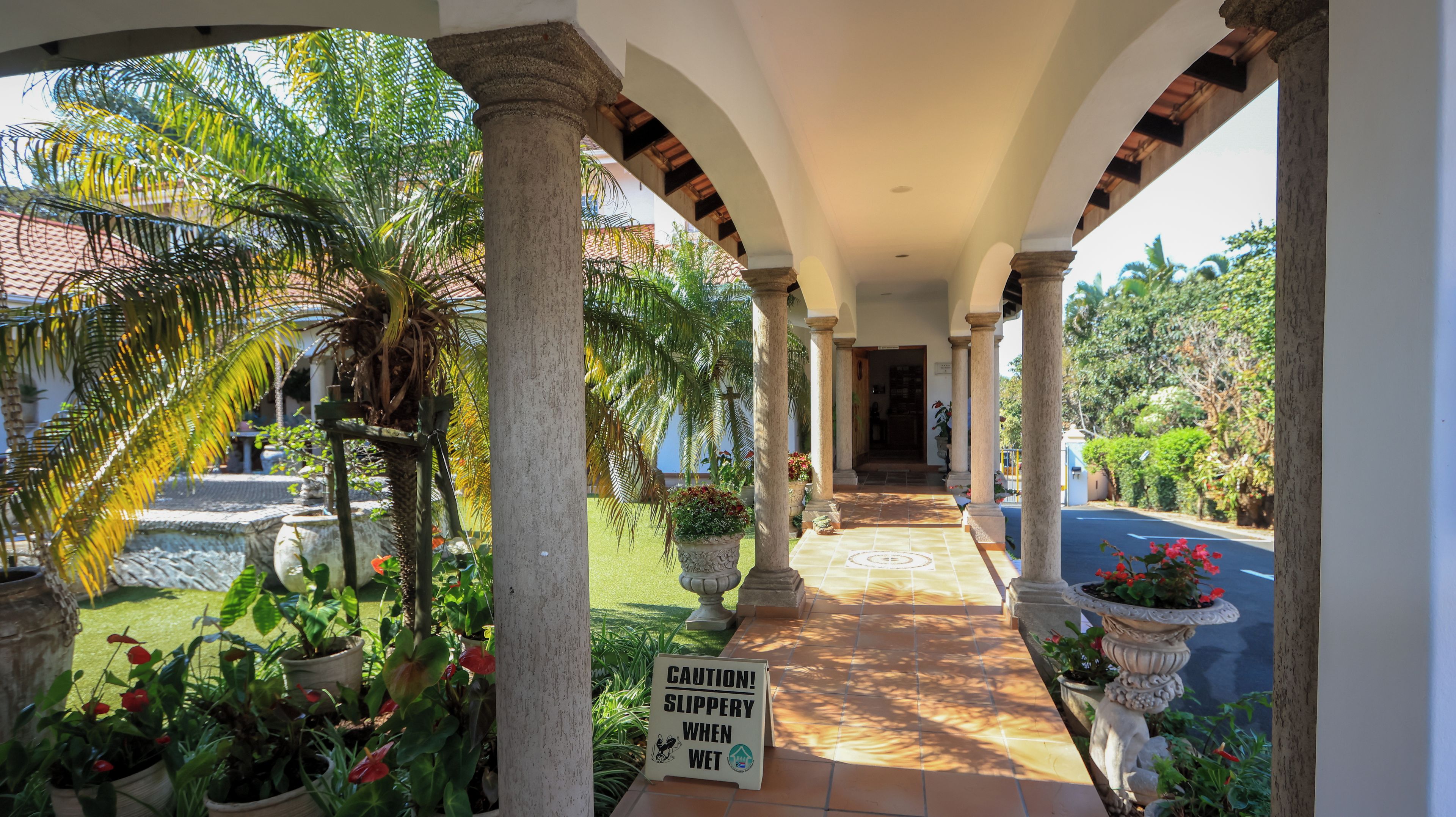 A covered outdoor corridor with columns, potted plants, and a caution sign.
