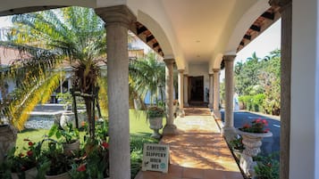 A covered outdoor corridor with columns, potted plants, and a caution sign.