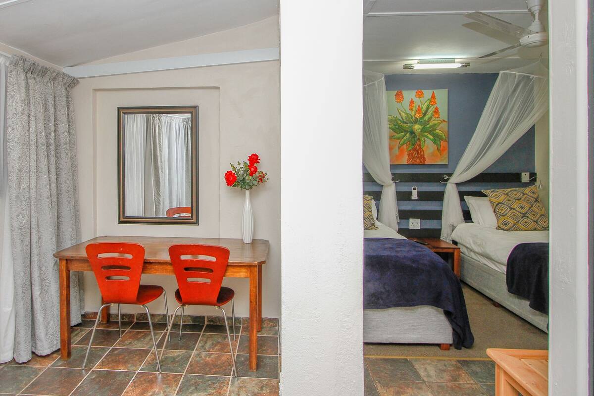 A room with a dining area featuring a wooden table and red chairs, a bed with a canopy, and a painting on the wall.