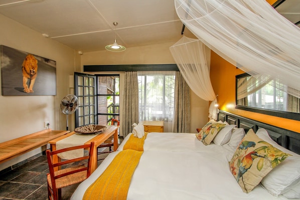 Habitación de hotel con cama, escritorio, silla, ventilador y ventana grande con cortinas.